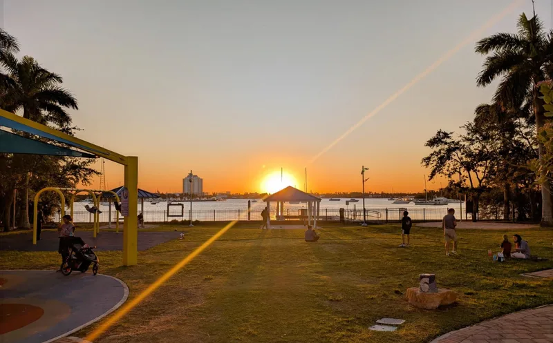 People enjoying a waterfront park at sunset, with a playground and pavilion beside the bay